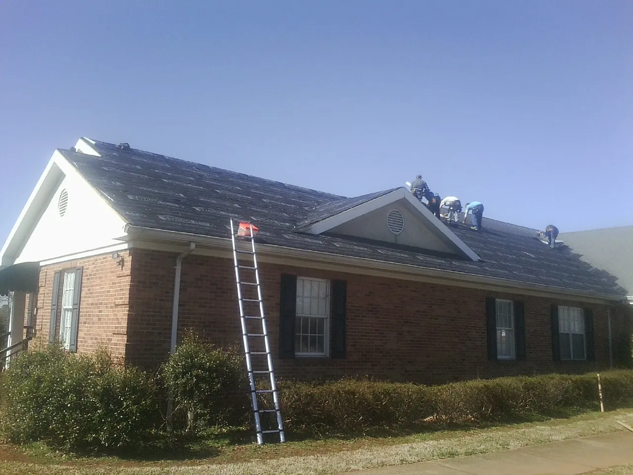 Crew tearing shingles off a brick ranch roof with a ladder set — Metal Roof Repair work by our crew in Orchard