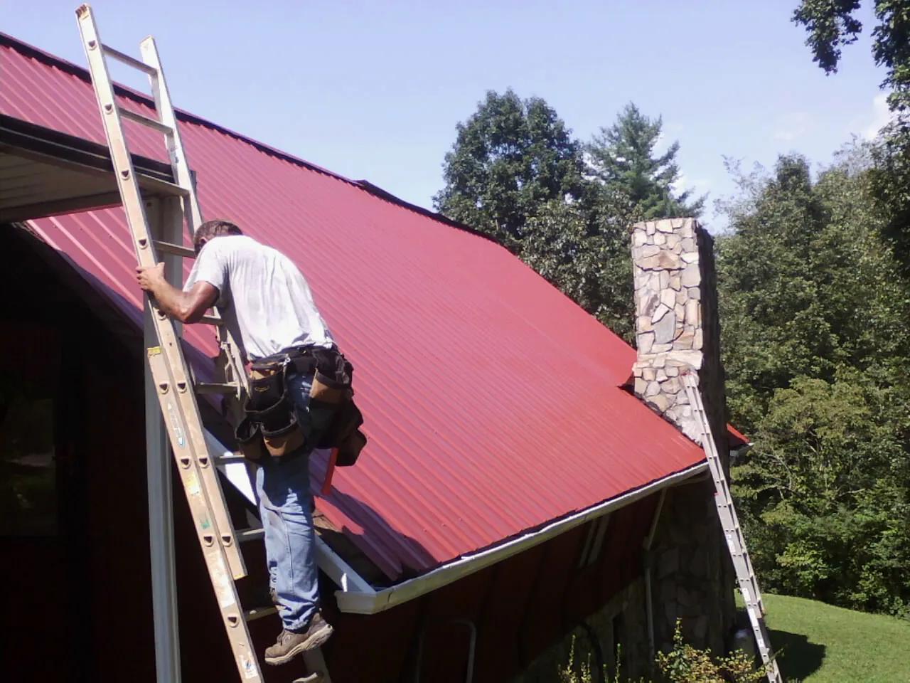 Roofer on a ladder beside a red metal roof and stone chimney — Soffit & Fascia Repair by Roofing Brothers in Orchard